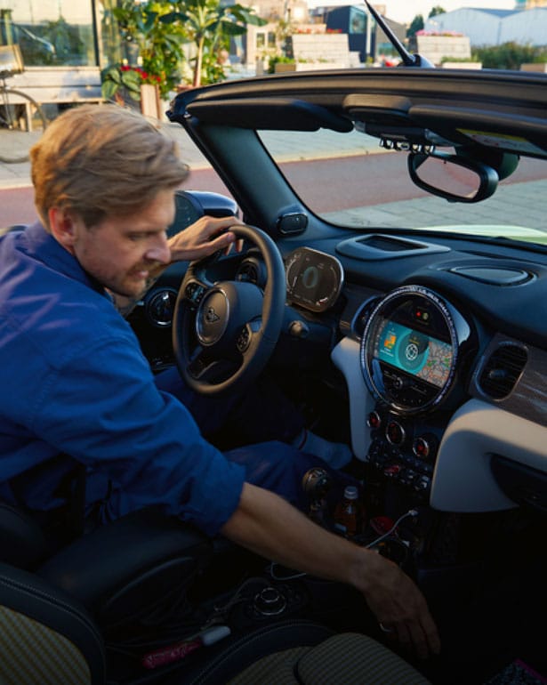 Man in blue longsleeve shirt reaching over onto the front passenger’s seat of a MINI Convertible, with the top down, while sitting in the driver’s seat.